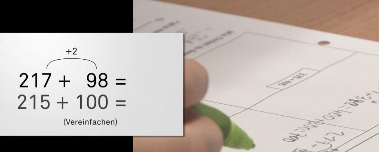 Ausschnitt aus dem PIKAS-Video zum Rechnen auf eigenen Wegen. Links: eingeblendet die Aufgabe „217 plus 98 = _“ und „215 plus 100 =_“. Darunter: „Vereinfachen“. Rechts: Nachaufnahme, wie ein Stift auf ein Arbeitsblatt schreibt.