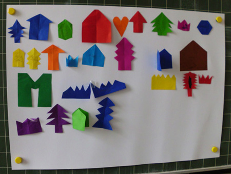 Foto eines Plakats mit Schülermaterial zu Faltschnitten. Darauf kleben verschiedene Faltschnitte aus buntem Papier. Zum Beispiel Tannenbäume, Häuser, Herzen.