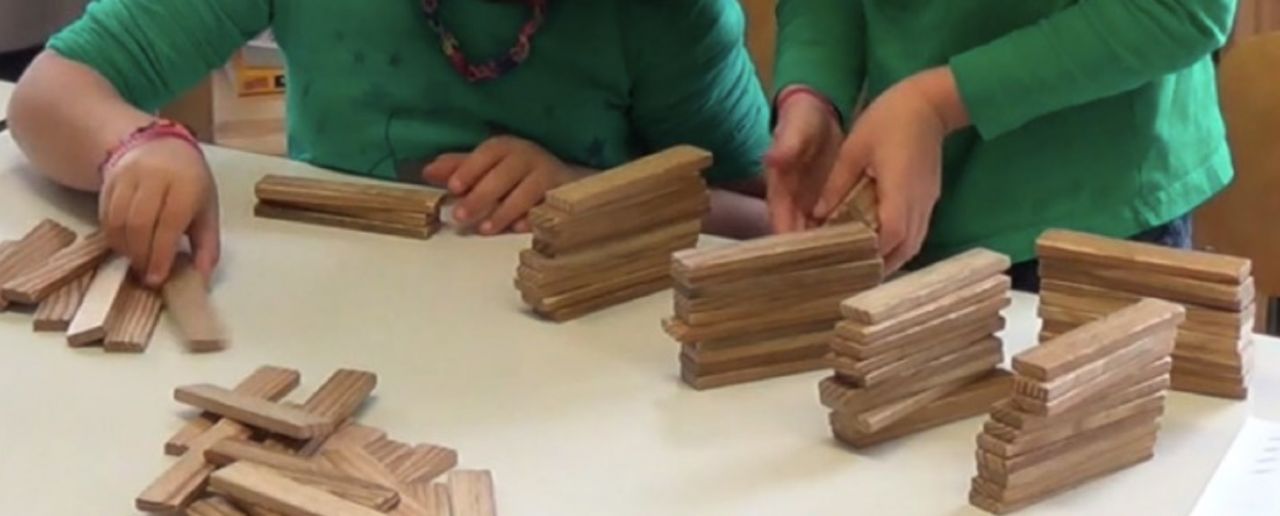 Ausschnitt eines Arbeitsplatzes von zwei Kindern. Sie hantieren mit Holzstäben. Rechts: Das Kind hat 5 Stapel mit jeweils 10 Holzstäben aufgebaut. Links: Das Kind hat einen Stapel mit 3 Holzstäben gebaut.